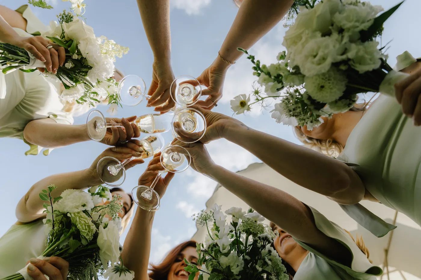 Bridesmaids holding bouquets raise champagne glasses in a toast, photographed from below against the sky
